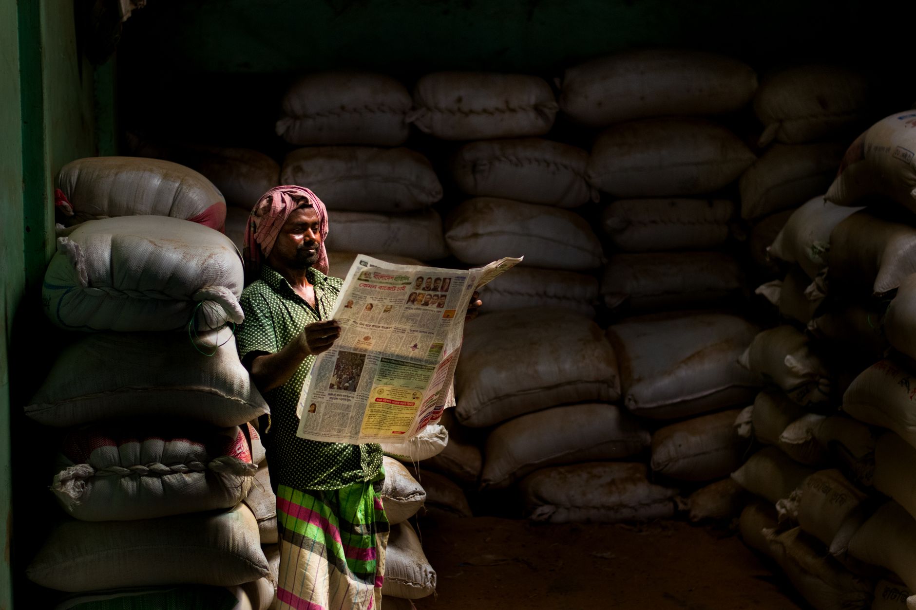 A man in traditional attire reads a newspaper in a dimly lit storeroom with stacked sacks. — Was macht Golden Pharaoh Casino